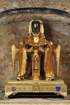 an ornate gold statue with angel wings on it's stand in front of a stone wall