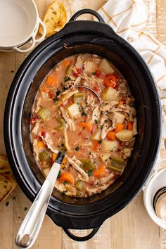 a crock pot filled with chicken and vegetable soup next to bread on a wooden table