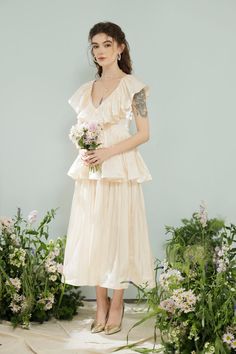 a woman standing in front of flowers and greenery wearing a dress with ruffles