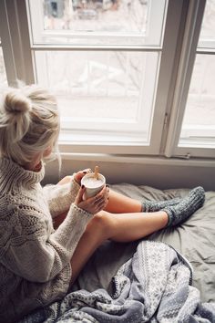 a woman sitting on top of a bed next to a window holding a cup of coffee