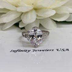 a diamond ring sitting on top of a table next to a white flower with the words infinity jewelers usa written below it