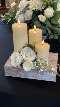 three candles are sitting on a wooden block with white flowers and greenery in the background