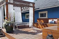 an outdoor hot tub on a wooden deck next to a blue house with white curtains