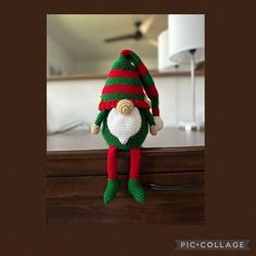 a green and red knitted elf doll sitting on top of a wooden dresser next to a lamp