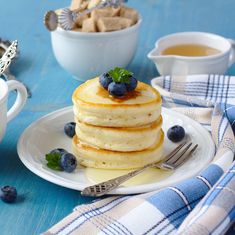 a stack of pancakes with blueberries and butter on a plate next to two cups of tea