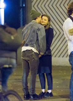 two people standing next to each other on a street corner with one person kissing the other