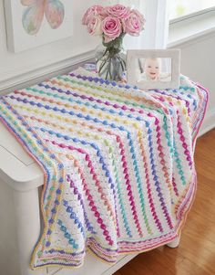 a crocheted afghan sits on a white bench next to a vase with pink roses