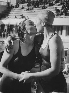 two women in bathing suits standing next to each other at a swimming pool, one kissing the other's cheek