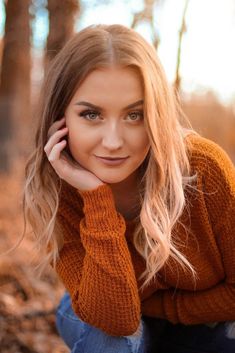 a beautiful blonde woman sitting in the woods with her hand on her head and looking at the camera