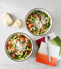 two bowls filled with pasta and vegetables on top of a white counter next to a piece of cheese