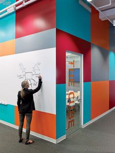 a woman writing on a whiteboard next to a wall with colorful walls and chairs