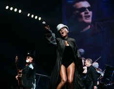 a woman in a black bodysuit and hat on stage with other performers behind her