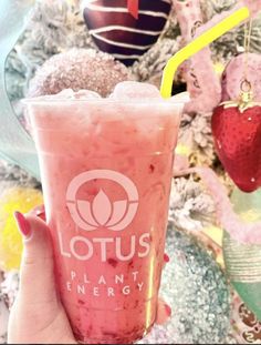 a person holding up a pink drink in front of a christmas tree with ornaments on it