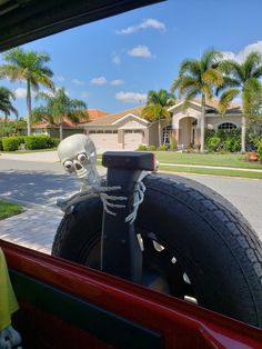 a skeleton sitting on the back of a red car in front of a house with palm trees