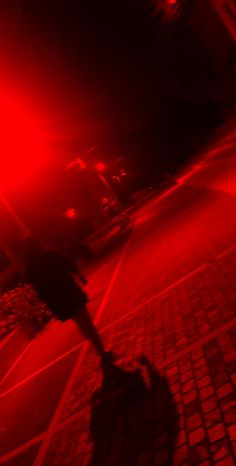 a person walking down the street at night with their shadow on the ground and red lights shining in the background