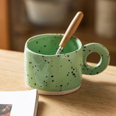 a green cup with a wooden spoon in it on a table next to a book