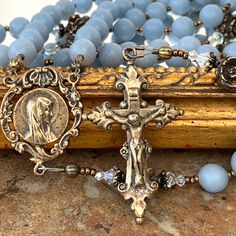 Artisan crafted featuring blue Angel Stone (Angelite) gemstones, Swarovski crystals and antique replicas cast in solid bronze using the lost wax method. The bronze crucifix was believed to originate from an early rosary-making town in Germany. The delicate scrolls on the ends of the crucifix pair nicely with the delicate scrolling and ribbon-style openwork found around the profile of the Our Lady Profile center. The Handsome Little crucifix measures 11/2.” The Profile center measures 11/8.” Each Spiritual Brass Miraculous Medal Jewelry, Spiritual Rosary With Miraculous Medal And Crucifix, Antique Bronze Crucifix Jewelry, Antique Finish Crucifix Jewelry Gift, Antique Crucifix Rosary Gift, Antique Crucifix Rosary As Gift, Vintage Bronze Rosary As Gift, Handmade Bronze Spiritual Rosary, Handmade Spiritual Bronze Rosary