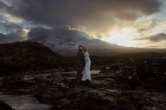 a man and woman standing on rocks in front of a mountain at sunset with the sun behind them