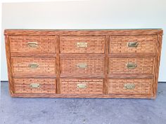 a wicker dresser with six drawers and brass pulls on the bottom drawer, in front of a white wall