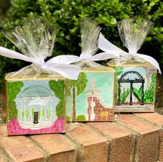 three small boxes with paintings on them sitting on top of a brick wall in front of some bushes