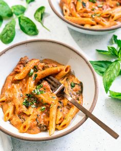 two bowls filled with pasta and sauce on top of a white table next to basil leaves