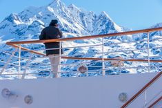 a man standing on the deck of a boat looking out at snow covered mountains in the distance