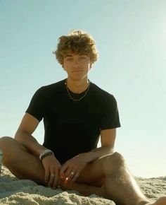 a young man sitting on the beach with his legs crossed and wearing jewelry, looking at the camera