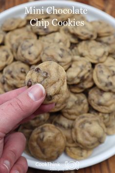 a hand holding a chocolate chip cookie in front of a white plate full of cookies