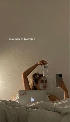 a woman laying in bed with an apple laptop and water bottle on her head while holding a cell phone