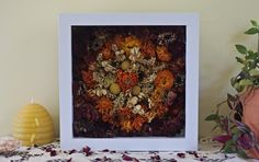 an arrangement of dried flowers in a white frame on a table next to a candle