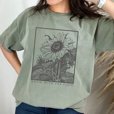 "This \"Grow With The Flow\" t-shirt is perfect for teachers, plant lovers, and anyone who wants to embrace the beauty of nature. The monochromatic sunflower design is pencil-drawn, giving it a vintage and retro feel. Made with Comfort Colors, this shirt is not only stylish, but also incredibly comfortable to wear. The botanical theme is perfect for garden and wildflower enthusiasts, and the floral tee makes a great addition to any wardrobe. Whether you're looking to inspire yourself or others, this shirt is a positive reminder to embrace the natural flow of life. Looking for a comfortable and durable t-shirt that you can customize to your liking? Look no further than Comfort Colors' garment-dyed tee! Here are some of the features that make this shirt a great addition to your wardrobe: -Ma Affordable Trendy Shirt With Plant Print, Green Cotton T-shirt With Plant Print, Cotton Graphic Print T-shirt For Gardening, Cotton T-shirt With Graphic Print For Gardening, Spring Crew Neck T-shirt For Outdoor, Green Cotton Shirt With Plant Print, Green Cotton Tops With Plant Print, Green Tops With Plant Print For Gardening, Spring Short Sleeve T-shirt For Gardening