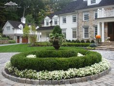 a circular garden in front of a large house with white flowers on the ground and bushes around it