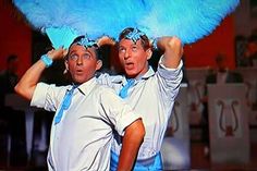 two men with blue feathers on their heads and one man holding up a large object