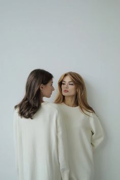 two women standing next to each other in front of a white wall and one is looking at the other