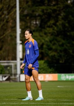 a man standing on top of a soccer field wearing blue and orange shorts with his hands in his pockets