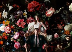 a man and woman are sitting on a chair in front of a large floral arrangement