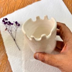 a hand holding a white vase on top of a paper towel next to a flower