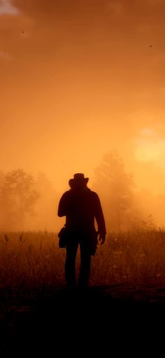 the silhouette of a man in a hat and coat walking through a field at sunset