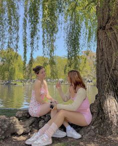 two young women sitting on the ground next to a tree and water, one holding a cell phone