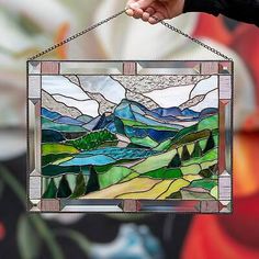 a hand holding a stained glass window with mountains in the background and clouds above it