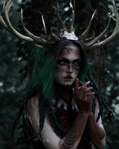 a woman with green hair and horns standing in front of some trees wearing antlers