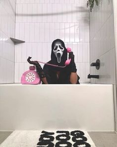 a person in a bathtub with a phone on the floor and a ghost mask