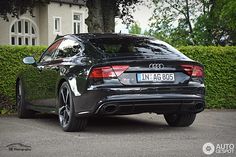 the rear end of a black car parked in front of a house with hedges behind it