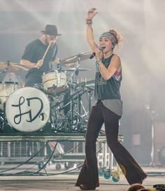 a woman on stage with her arms in the air and two drummers behind her