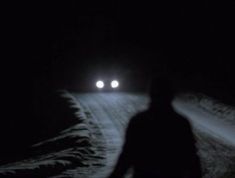 two people walking in the snow at night with their headlights turned on and one person standing next to them