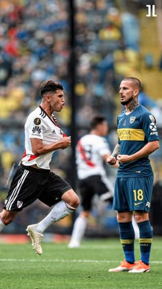 two men playing soccer on a field with fans in the stands and one man running after the ball