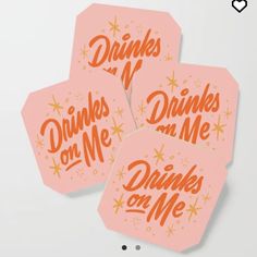 three pink drink coasters with drinks on me written in orange and gold lettering, against a white background