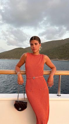 a woman in an orange dress leaning on a railing next to the ocean with her purse