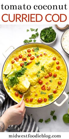 a person holding a spoon in a bowl filled with yellow curry and topped with tomatoes