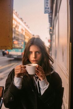 a woman holding a coffee cup in front of her face while looking out the window